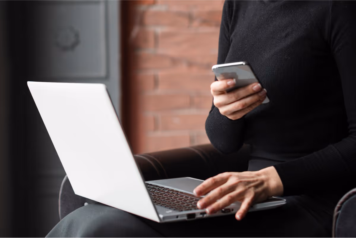 Person using a laptop and holding a smartphone while sitting in a chair.